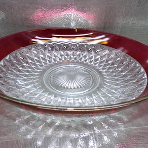May include: A clear glass serving bowl with a ruby red rim and a diamond-cut pattern. The bowl has a gold-colored trim and a circular design in the center. This decorative bowl is suitable for serving salads or fruit.