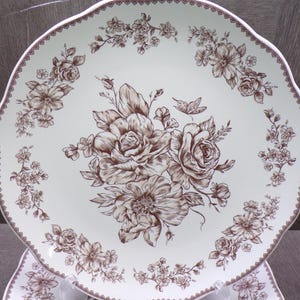 May include: A round ceramic plate with a scalloped edge featuring a cream-colored base and a brown floral transfer pattern. The design includes a central bouquet of roses and smaller floral sprigs arranged around the rim with a lattice border.