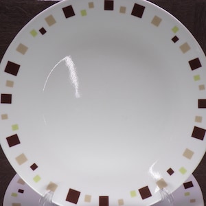 May include: White ceramic plate with a square rim design. The rim features alternating brown, beige, and light green squares. The plate is round with slightly curved edges, and is displayed on a clear plastic stand.