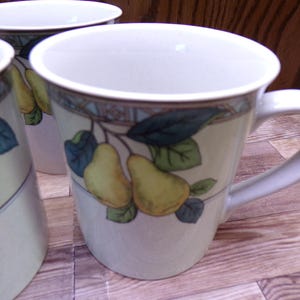 May include: White ceramic mugs with a pear and leaf design. The mugs have a handle and a decorative border. The mugs are on a wooden surface. The mugs are part of a set.