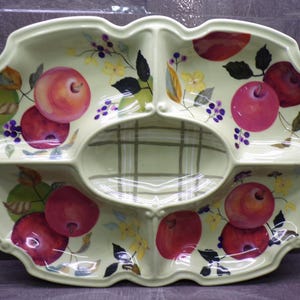 May include: A pale green ceramic serving dish with four outer compartments and one central oval section featuring a painted pattern of red apples, dark leaves, and small purple berries. The central section has a light green grid pattern.
