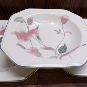 May include: Three white octagonal soup bowls with a floral design featuring pink flowers, green leaves, and brown stems. The bowls have a blue trim and are arranged on a wooden surface.