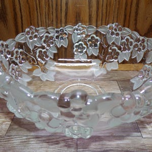 May include: A clear glass bowl with a floral design. The bowl has a wide, oval shape with a frosted finish and detailed relief of flowers and leaves around the rim. The flowers have a brown center.