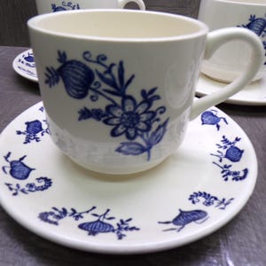 May include: Cream-colored teacups and saucers with a blue floral design. The cups have a curved handle and sit on matching saucers. The blue pattern includes flowers and other botanical elements. The set is likely porcelain.