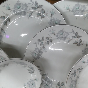 May include: A set of five white ceramic plates with a silver trim and a grey floral design. The plates are arranged in a stack, with the largest plate at the bottom and the smallest plate at the top.