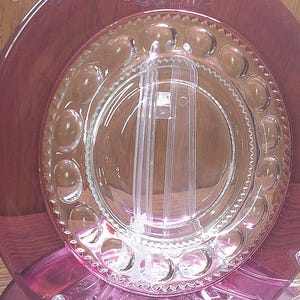 May include: A clear glass plate with a raised rim and a pattern of small circles around the edge. The plate is sitting on top of a larger, pink glass plate.