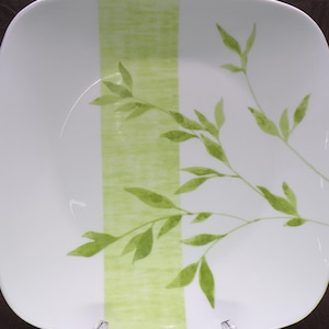 May include: Square white plate featuring a light green stripe and a green leaf and branch design. The plate has rounded corners and a glossy finish, suitable for serving food. The plate is approximately 20 cm square.