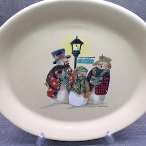 May include: An oval cream-colored ceramic platter featuring a painted illustration of three snowmen standing under a street lamp. The snowmen wear hats, scarves, and patterned coats. A sign labeled N PENNY LN hangs from the lamp post. The artist signature is visible at the bottom right.