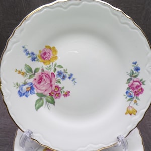 May include: White porcelain plate with a scalloped edge and gold trim. The plate features a floral design with pink roses, yellow and blue flowers, and green leaves. The plate is displayed on a clear stand.