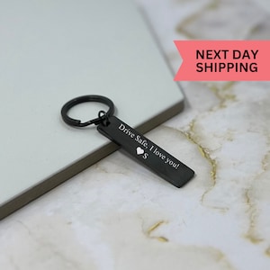 Peut inclure: Porte-clés en métal noir avec un anneau argenté et une étiquette rectangulaire sur laquelle est inscrit "Drive Safe, I love you!" avec un symbole de cœur.