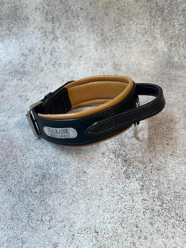 May include: Black and tan leather dog collar with a padded interior. The collar has a silver-colored buckle and a small metal tag with the name "VALKANE" and a phone number. The collar is designed for comfort and durability.