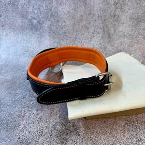 May include: A black leather dog collar with an orange interior. The collar has a silver-colored metal buckle and a D-ring for leash attachment. The collar is resting on a white canvas bag.