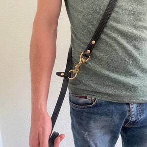 May include: A brown leather dog lead with a gold metal clip and brass stud detail. The lead is worn over the shoulder of a person wearing a green t-shirt and blue jeans.