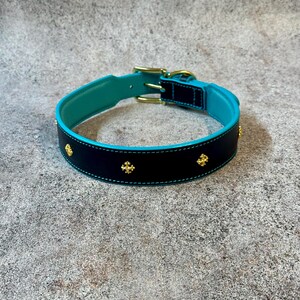 May include: A black dog collar with a turquoise interior and trim, adorned with gold-colored cross-shaped embellishments. The collar features a gold-colored buckle and hardware. The stitching is a light turquoise color.
