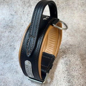 May include: A black and tan leather dog collar with a handle and a metal D-ring. The collar has a padded interior and a metal nameplate with the name "VALKANE" and a phone number. The collar is designed for durability and comfort.