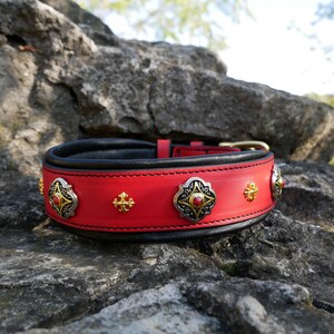 May include: A red leather dog collar with black lining and gold-coloured hardware. The collar is adorned with silver and gold-coloured decorative elements, including ornate medallions with red centre stones and smaller gold crosses.