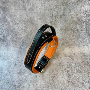 May include: A black and orange leather dog collar with a handle. The collar features a silver-colored buckle and a D-ring for leash attachment. The collar is made of black leather with orange padding and stitching.