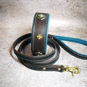 Pu&ograve; includere: Un set di collare e guinzaglio per cani in pelle marrone con dettagli e cuciture blu turchese. Il collare presenta decorazioni decorative in metallo argentato e dorato. Il guinzaglio ha un moschettone color ottone.