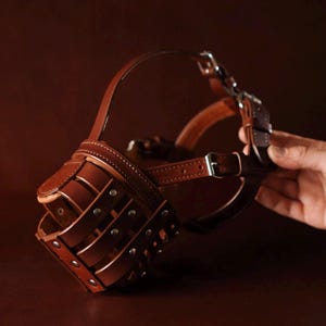 May include: A brown leather dog muzzle featuring a basket-style cage design with multiple horizontal and vertical straps. The muzzle is secured with silver-toned metal rivets and includes adjustable leather straps with metal buckles for fastening.