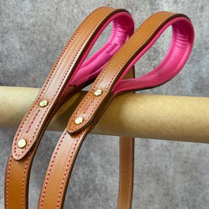May include: Two tan leather leashes featuring bright pink padded handles and pink stitching. Each leash is secured with gold-toned metal rivets at the handle junction, showcasing a smooth leather finish and a vibrant, contrasting interior lining.