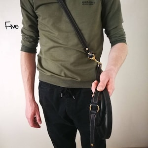 May include: Black leather dog leash with brass-colored hardware. The leash is held by a person wearing a green long-sleeve shirt and black pants. The leash has a stitched design.