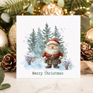 May include: A square greeting card featuring a watercolour illustration of a smiling Santa Claus wearing a red coat and a plaid kilt, standing in a snowy forest with pine trees. The text Merry Christmas is printed in dark green at the bottom.