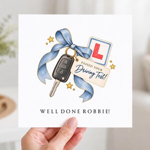 Peut inclure: Une carte de vœux carrée avec une illustration à l'aquarelle d'une clé de voiture noire attachée par un ruban bleu, un panneau L de conducteur novice et une étiquette indiquant PASSED YOUR DRIVING TEST. WELL DONE ROBBIE est inscrit en bas.