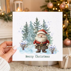 May include: A square greeting card featuring a watercolour illustration of a smiling Santa Claus wearing a red jacket and a plaid kilt, standing in a snowy forest with pine trees. The text Merry Christmas is printed in black at the bottom.