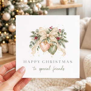 May include: A square greeting card featuring a watercolour illustration of two heart-shaped ornaments in pink and green, accented with a large beige ribbon, pine branches, and red berries. The text reads: HAPPY CHRISTMAS to special friends.