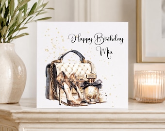 Op de afbeelding: Een vierkante wenskaart met een witte gewatteerde handtas, een paar sandalen met hoge hakken met een gouden glitterafwerking en een parfumflesje met een zwarte strik. De tekst Happy Birthday Mia staat in zwarte letters boven de items.