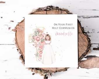 May include: A white card with the text "ON YOUR FIRST HOLY COMMUNION" and the name "Annabelle" in pink script. The card features a girl in a white dress and a gold cross adorned with pink roses and greenery.