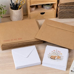 May include: A stack of rectangular brown kraft paper envelopes with the red text PLEASE DO NOT BEND printed on the front, alongside a stack of square white envelopes and a stack of square white greeting cards featuring a sleeping fox illustration.