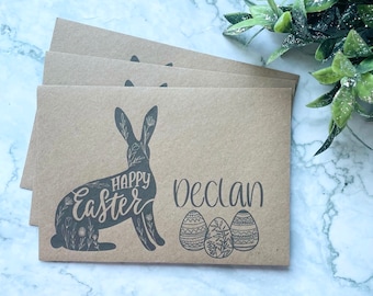 May include: Kraft paper Easter cards with a black floral bunny design and the words "Happy Easter" and "Declan". Three decorated Easter eggs are also featured. The cards are stacked on a white and grey marble surface.