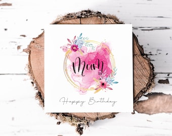 May include: A white card with a pink watercolour background and gold glitter. The word "Mum" is written in black cursive script inside a gold circle. The card also has a floral design with pink, blue, and yellow flowers. The text "Happy Birthday" is written in black cursive script at the bottom of the card.