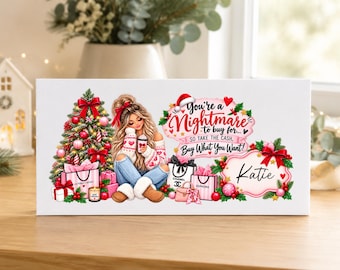 Puede incluir: Un letrero rectangular blanco con una ilustración festiva de una persona de cabello largo sentada junto a un árbol de Navidad decorado, rodeada de bolsas de compras y regalos. El texto dice: You're a Nightmare to buy for... SO TAKE THE CASH, Buy What You Want! El nombre Katie está escrito en un marco decorativo.