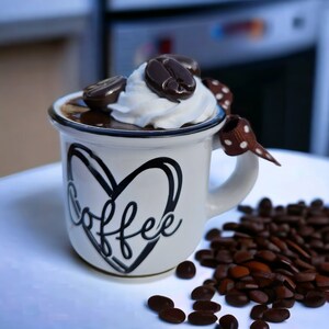 May include: White ceramic coffee mug with a black heart and the word "coffee" written inside. The mug is filled with chocolate pudding and topped with whipped cream and chocolate coffee beans. The mug is sitting on a white surface with coffee beans scattered around it.