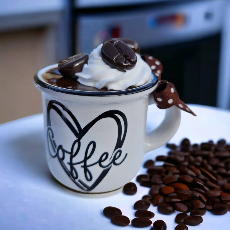 May include: White ceramic coffee mug with a black heart and the word "coffee" written inside. The mug is filled with chocolate pudding and topped with whipped cream and chocolate coffee beans. The mug is sitting on a white surface with coffee beans scattered around it.