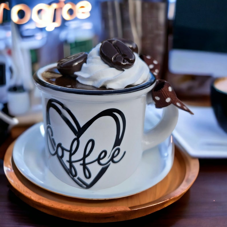 May include: White ceramic coffee mug with a black heart and the word "coffee" written inside. The mug is filled with a brown liquid topped with whipped cream and chocolate pieces.