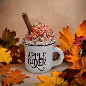 May include: White ceramic mug with the words "Apple Cider" in black lettering. The mug is filled with a brown and white whipped cream topping with cinnamon sticks and red and orange sprinkles.