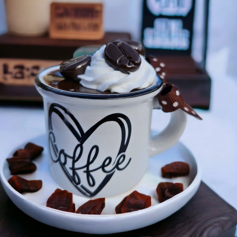 May include: White ceramic coffee mug with a black heart and the word "coffee" written inside. The mug has a brown and white polka dot ribbon tied to the handle. The mug is filled with chocolate mousse topped with whipped cream and chocolate coffee beans.