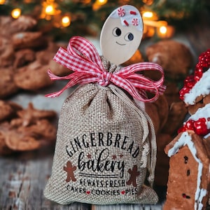 May include: A burlap bag with the text "Gingerbread Bakery" and a gingerbread man graphic. A wooden spoon decorated as a gingerbread person is tied to the bag with a red and white checkered ribbon. The background features cookies and Christmas lights.