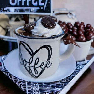 May include: A white coffee mug with a black heart and the word "coffee" written inside. The mug is filled with a chocolate-colored liquid topped with whipped cream and a chocolate coffee bean.