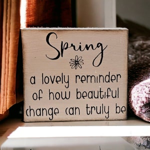 May include: A white wooden sign with black lettering that reads "Spring, a lovely reminder of how beautiful change can truly be". A small flower graphic is above the word "Spring".