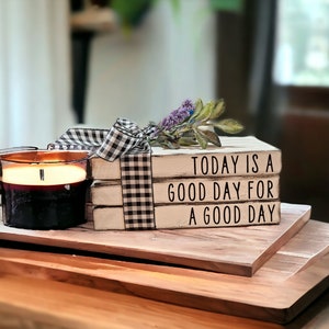 May include: Three white wooden blocks stacked on top of each other with black lettering. The top block reads "TODAY IS A", the middle block reads "GOOD DAY FOR", and the bottom block reads "A GOOD DAY". The blocks are decorated with a black and white checkered ribbon and a sprig of purple flowers.