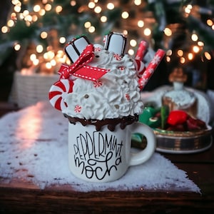 May include: White ceramic mug with a red and white peppermint mocha design. The mug is topped with a white whipped cream and red and white candy.