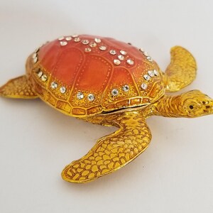 Peut inclure: Une petite boîte à bijoux en forme de tortue de couleur dorée avec une carapace rouge décorée de strass. La tête de la tortue est levée et ses pattes sont tendues.