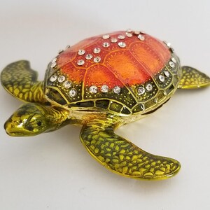 Peut inclure: Une petite figurine de tortue orange et verte avec des strass sur sa carapace. La tortue est en métal et présente un design détaillé.