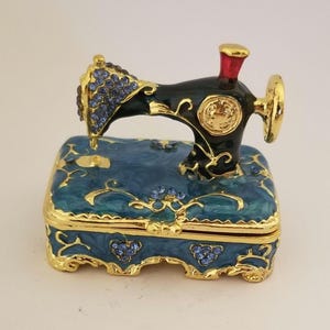 Peut inclure: Une boîte à bijoux en émail bleu et or en forme de machine à coudre vintage. La machine est décorée de strass bleus et a une base dorée.