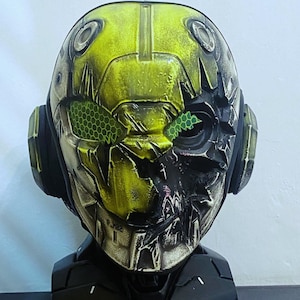 May include: A yellow and black robot helmet with a cracked and weathered finish. The helmet has a green honeycomb design on the eyes and a black base.