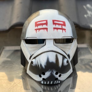 May include: A white and gray helmet with a black visor and red markings. The helmet has a stylized design with a mouth and teeth.